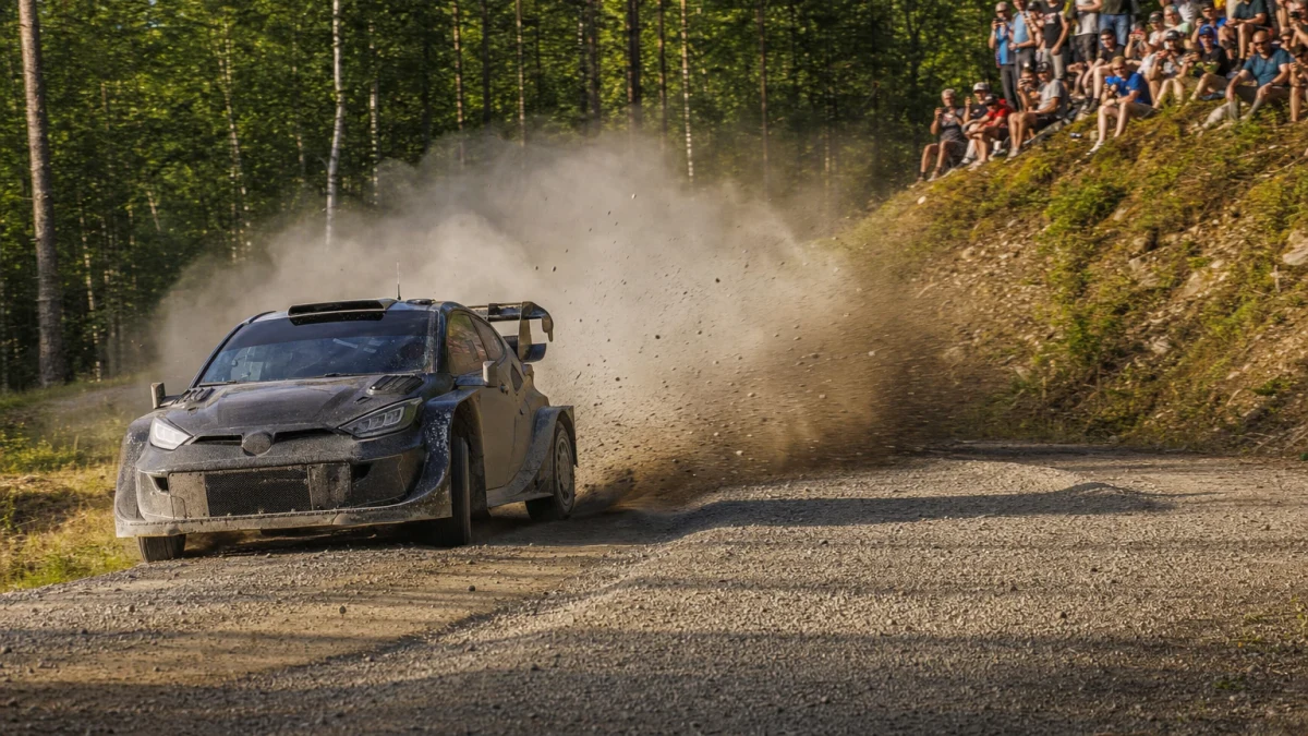 Illustration: rallybil som kastar upp grus och damm genom en kurva på en grusväg i skogen, med åskådare på slänten bakom.